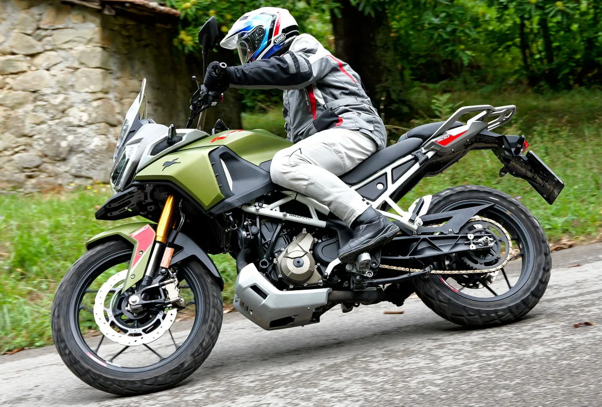 Une moto TVS RTX 300 penchée en roulant sur une route, un motard en tenue trail grise et un casque bleu blanc rouge Shoei Hornet.