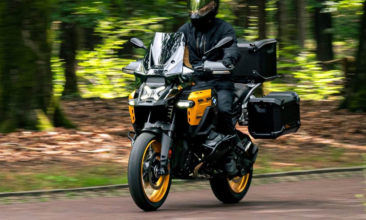 Une moto BMW R 1300 GS Adventure Aurum Edition jaune avec des valises et un top-case roulant sur des pavés et une forêt.