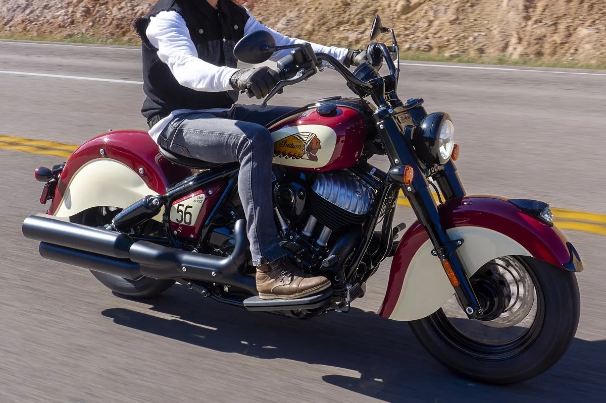 Une moto Indian 1890 Chief Vintage Sturgis, SD Edition, coloris Red & Cream, un potard avec un jean, un gilet et un pull blanc, une route et une bande jaune