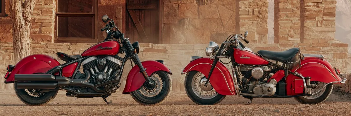 Une Indian chief vintage de 2026 couleur bordeaux à coté d'une indian Chief de 1940, sur un sol poussiéreux devant une vieille bâtisse