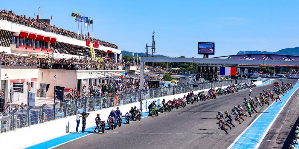 Le depart d'une course moto, des pilotes en train de courir, une ligne droite de circuit, des motos et des gradins remplis de spectateurs