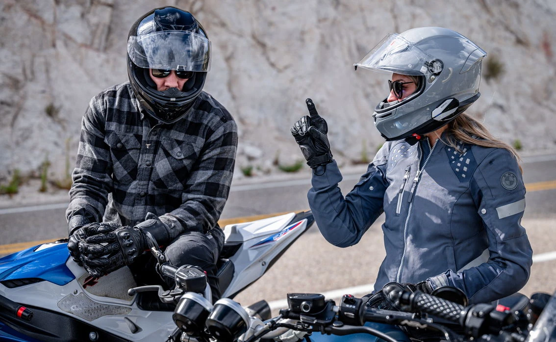 Un motard avec un casque noir et une veste à carreaux gris et noirs et Une motarde avec un casque gris et le doigt levé dans un gant noir.