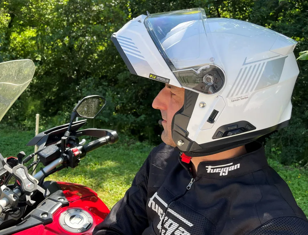 Un casque moto modulable Sena Specter Blanc en position ouverte, un réservoir de moto rouge, de la végétation, un blouson Furygan