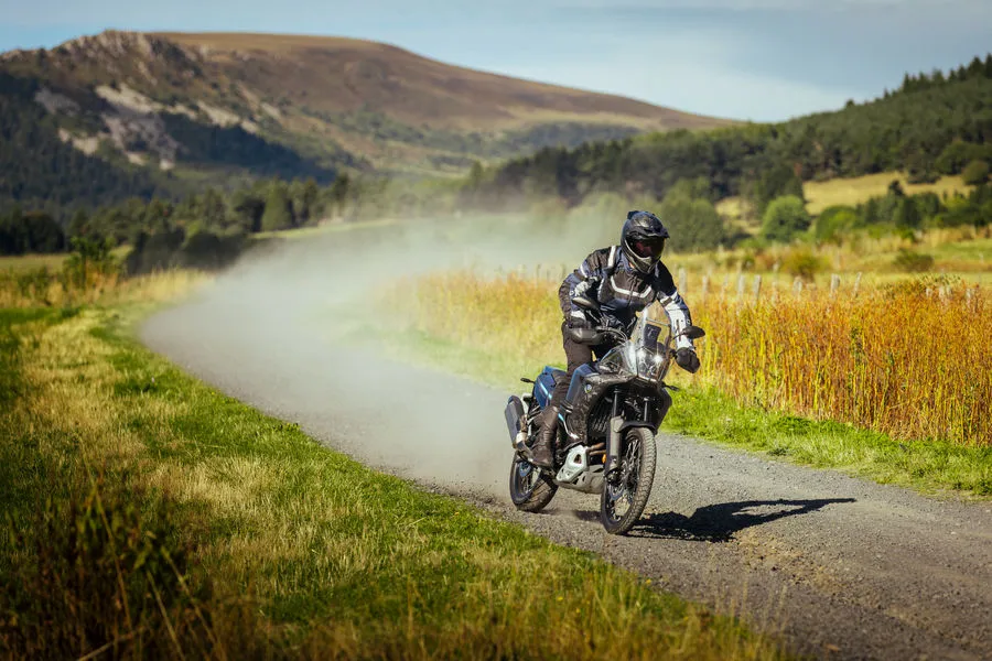Une Yamaha 700 Ténéré roulant sur un chemin de terre avec des herbes hautes et une colline dans le fond