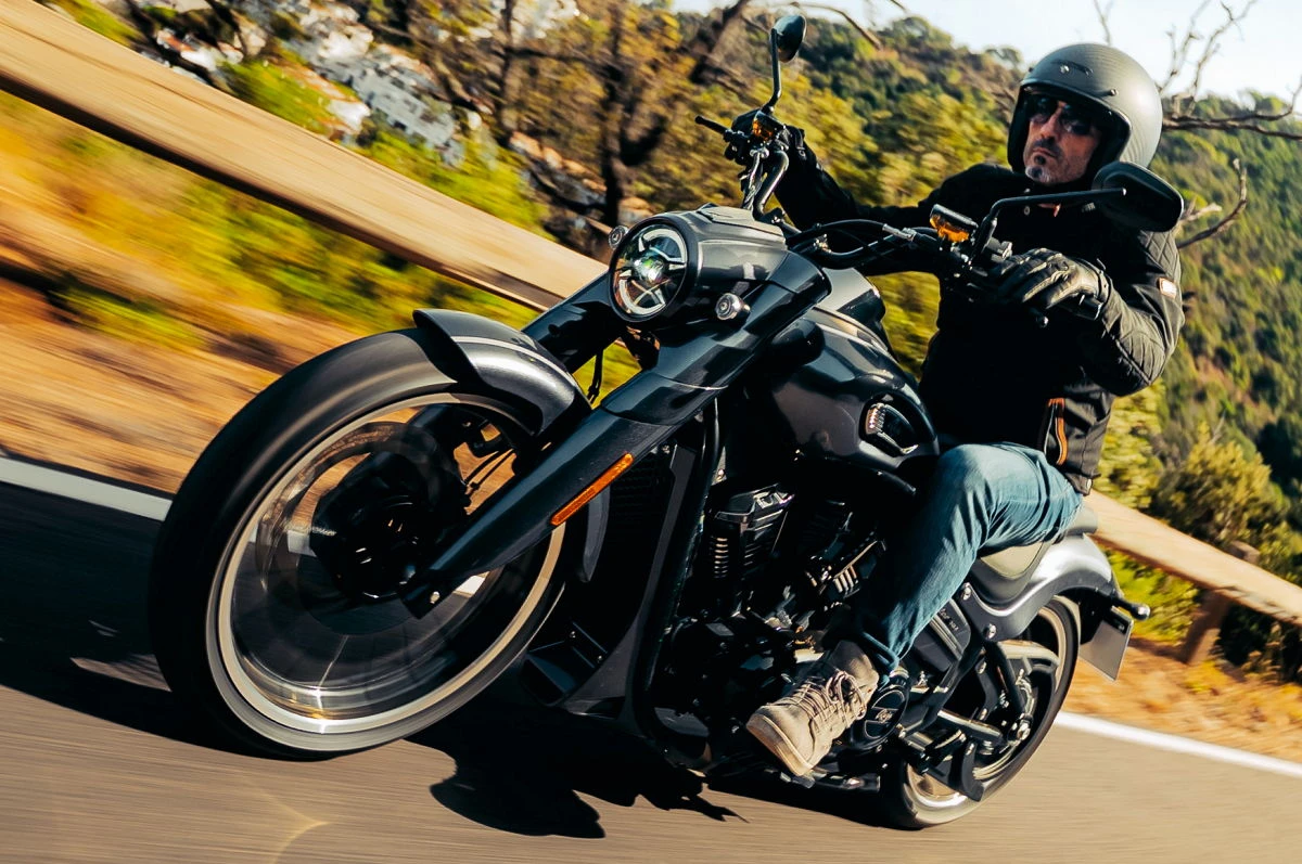Un custom Rieju Coaster 407, une route, des arbres, un motard en jean et blouson noir et casque jet.